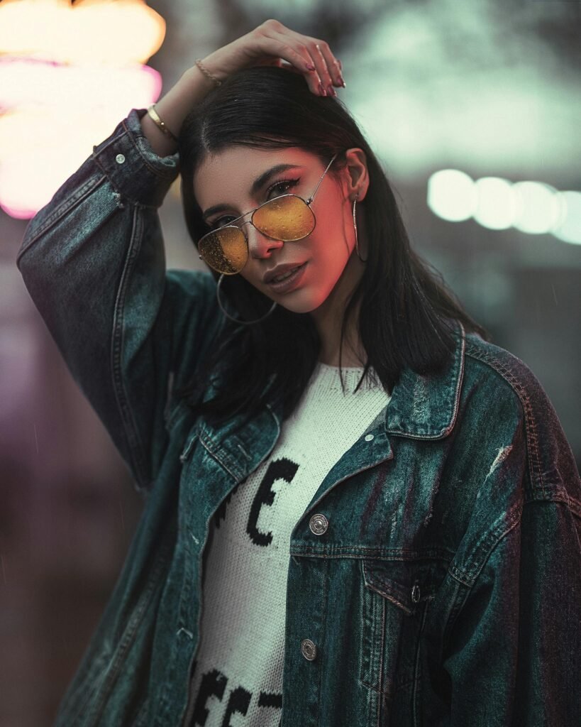 Fashionable woman posing confidently with sunglasses and denim jacket during a night shoot.
